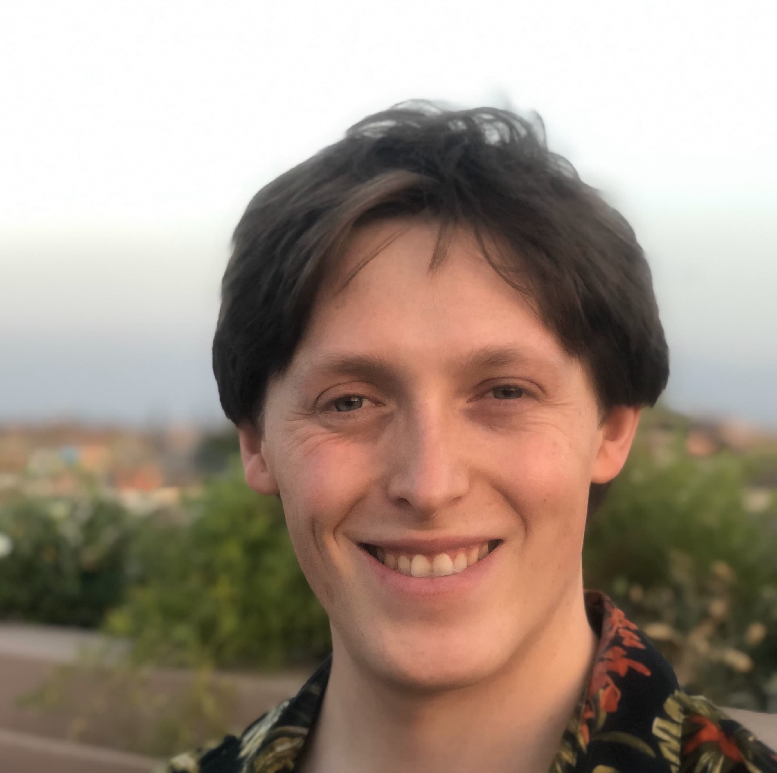 A photo of Dylan, taken in 2019 from a rooftop restaurant in Morocco. Pictured with medium length brown hair, slightly disheveled and parted on the viewer's right, is a wide-smiling, green-eye squinting Dylan wearing a black floral-print collared shirt. The image is neck-up, and the shirt is barely visible in the crop. A portrait style photo, the head makes way to slightly blurred shrubbery, offering a verdant contrast to the subject's peach-toned skin. Further in the photo's background we see the Old Town of Morocco stretch to the horizon, beige and reddish-brown buildings carrying our eyes to the gray-blue sky behind up.